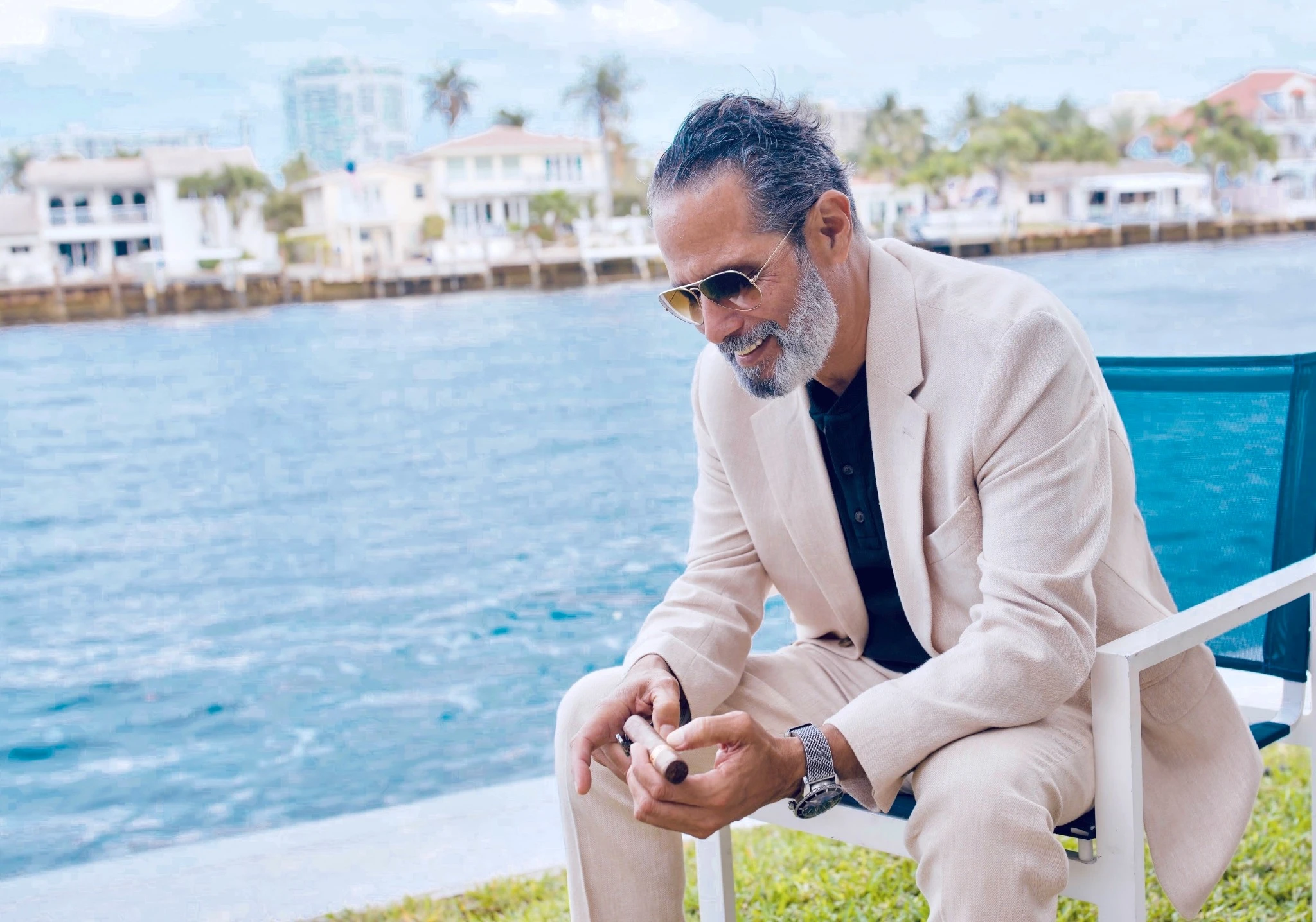 Anthony in a cream suit, seated by the water holding and cutting a cigar